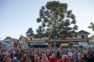 Pessoas aplaudem uma orquestra em um palco ao ar livre, com o sol se pondo e árvores ao fundo.